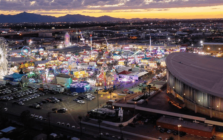 Aerial view of the Arizona State Fair in Phoenix at dusk, showing bright carnival lights, rides, food stands, and the surrounding cityscape against a sunset sky.