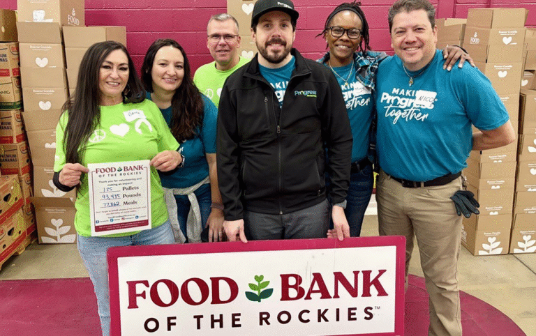 A group of volunteers standing together at the Food Bank of the Rockies, surrounded by stacked boxes, with one person holding a sign displaying completed donation totals.